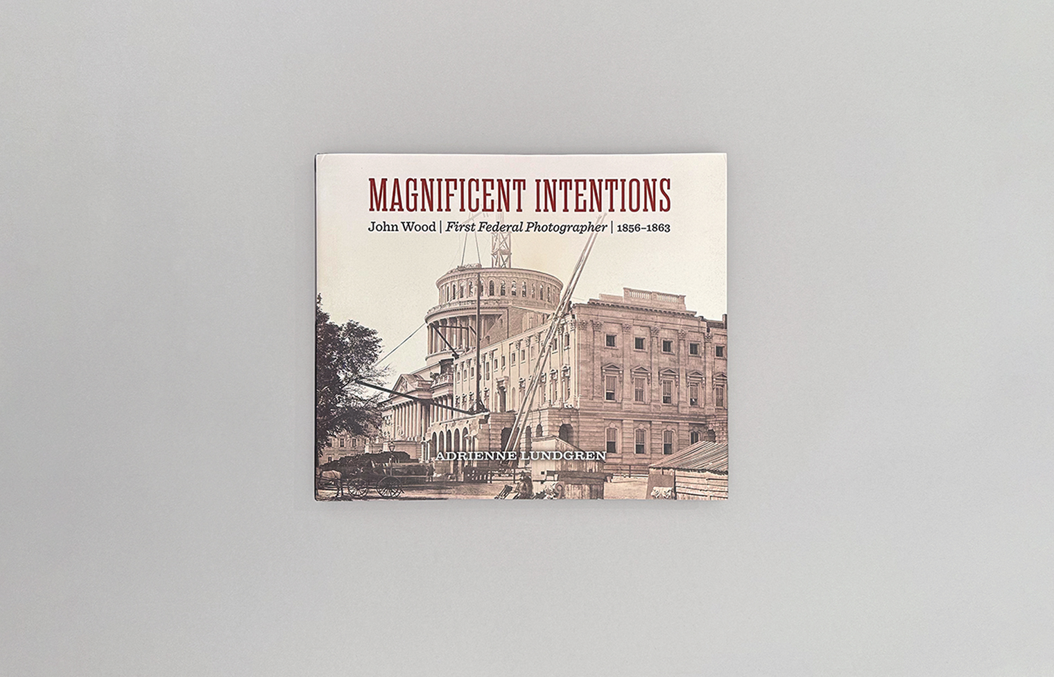 The image shows the cover of a book titled Magnificent Intentions: John Wood | First Federal Photographer | 1860–1865 by Adrienne Lundgren. The cover features a sepia-toned photograph of the U.S. Capitol building under construction with scaffolding, set against a gray background.