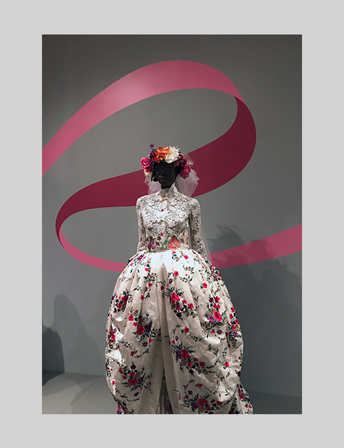 Exhibition photo from Inspiring Beauty: 50 Years of Ebony Fashion Fair shows a white floral dress with a ribbon wall graphic.