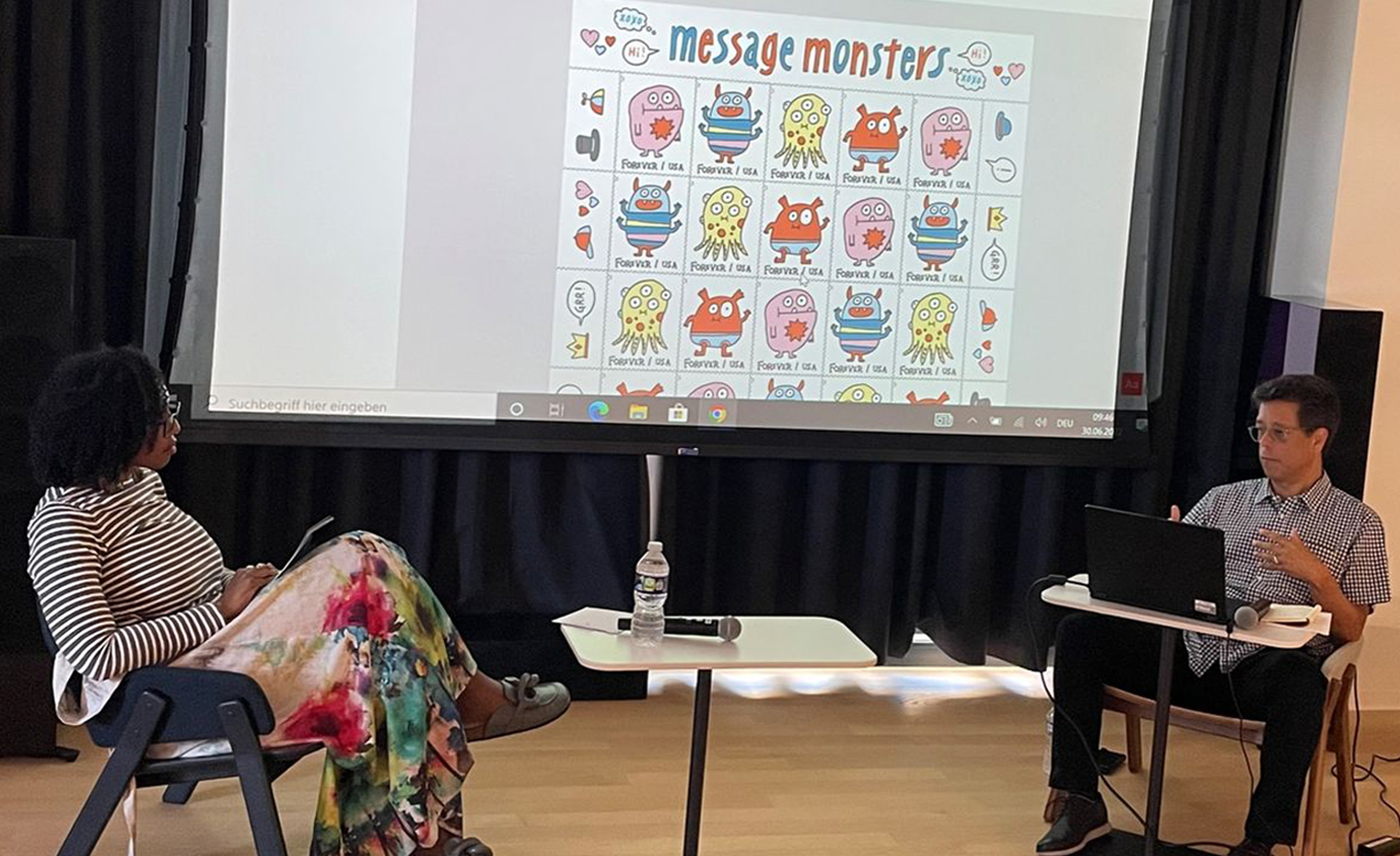 Dian Holton and Antonio Alcala seated in front of a screen that has the Message Monsters stamp on screen. It features whimsical creatures and additional stickers that can be added by the sender on the stamp